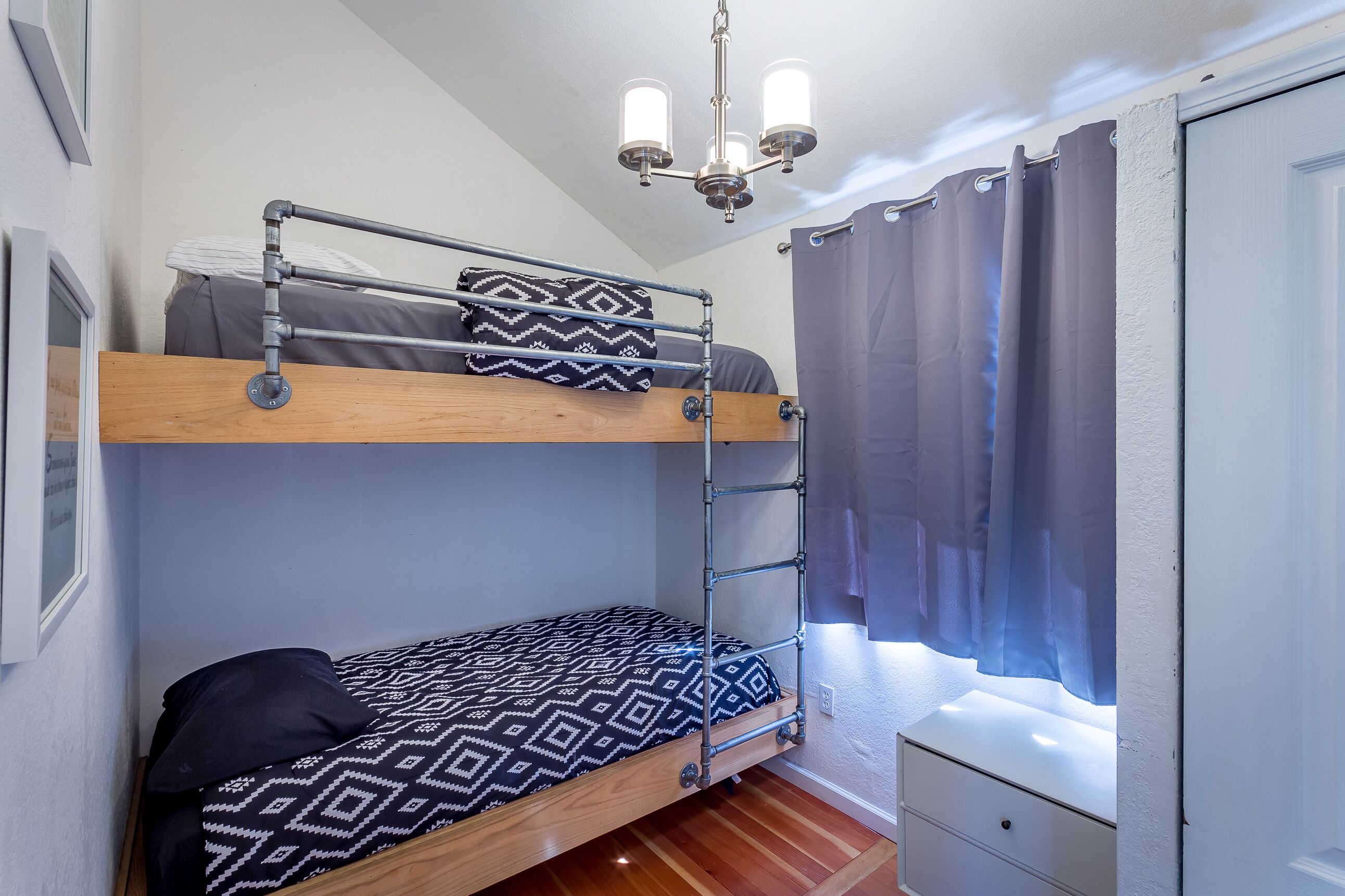 A bunk bed with a ladder, a bedside table, a window with curtains, and a chandelier.