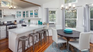 A modern kitchen with white cabinets, a central island with stools, a dining table with chairs, and large windows with curtains.