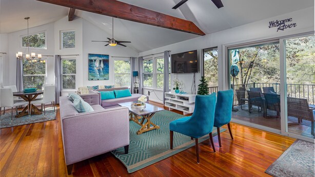 A spacious living room with a large sofa, a coffee table, and a TV. There are multiple windows and a balcony with outdoor furniture.