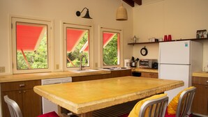 Eine kleine Küche mit einem Holztisch, roten und gelben Sitzkissenstühlen, einem weißen Kühlschrank und einem Fenster mit roter Markise.