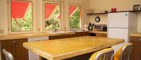 Une cuisine de taille modeste, avec une table en bois, des chaises rembourrées aux couleurs rouge et jaune, un réfrigérateur blanc et une fenêtre surmontée d’un auvent rouge.