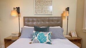 A neatly made bed with a headboard, two bedside lamps, a framed picture, and a small table with a lamp and a box.