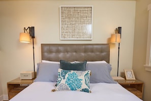 A neatly made bed with a headboard, two bedside lamps, a framed picture, and a small table with a lamp and a box.