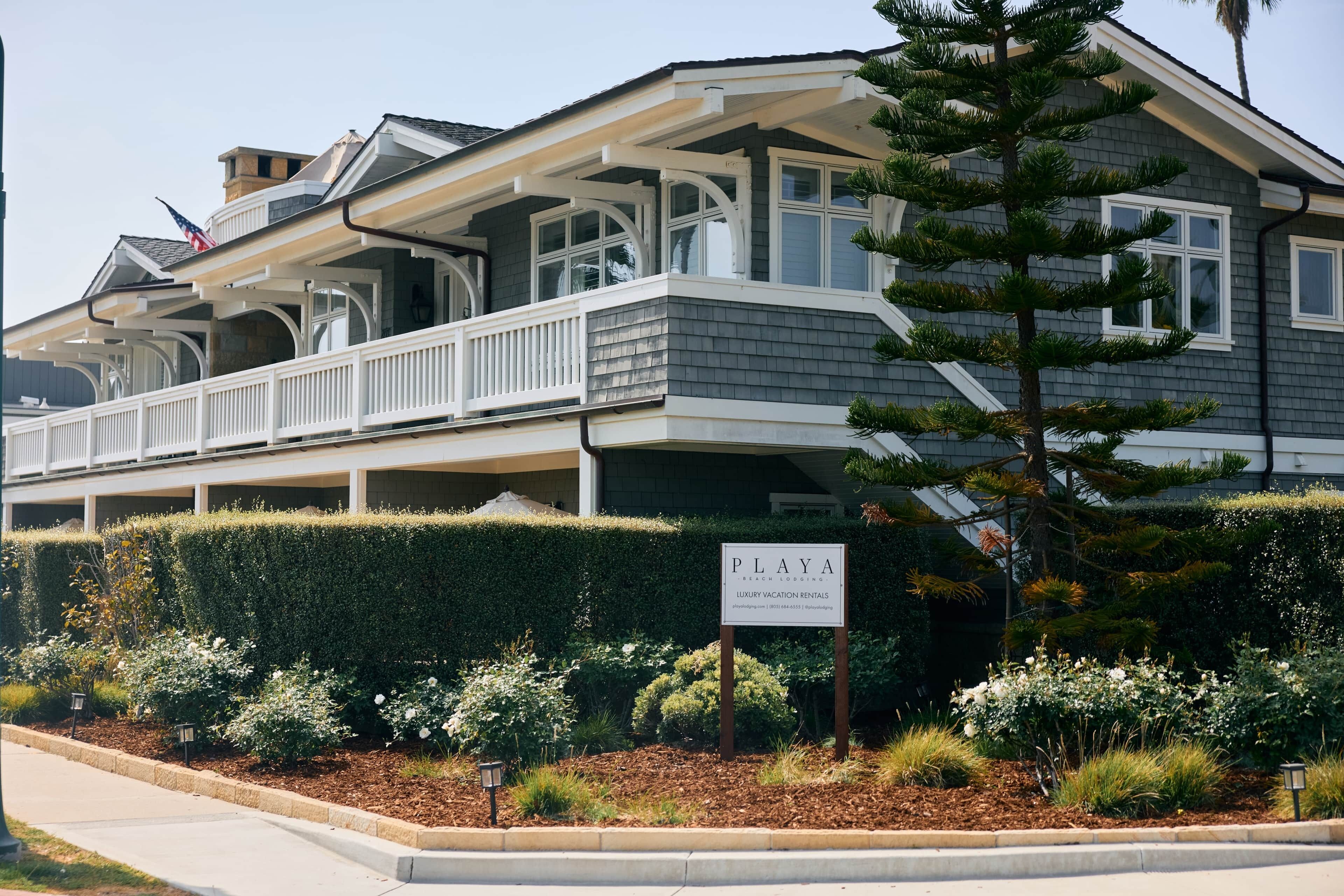 A two-story building with a balcony, a sign indicating it is a beach lodging, and a well-maintained garden with various plants and flowers.