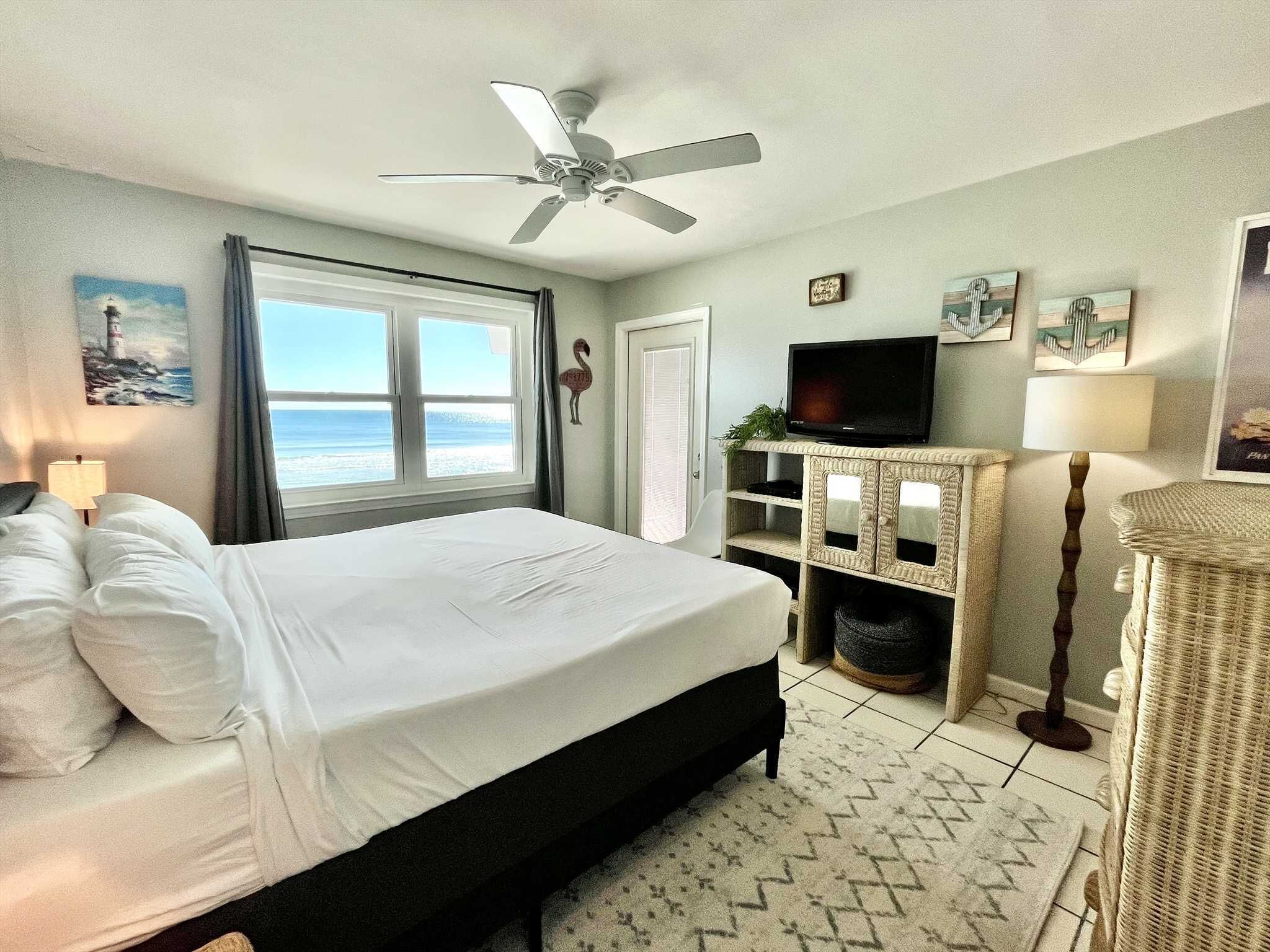 A hotel room with a bed, a television, a ceiling fan, a window with a view of the sea, and a door.