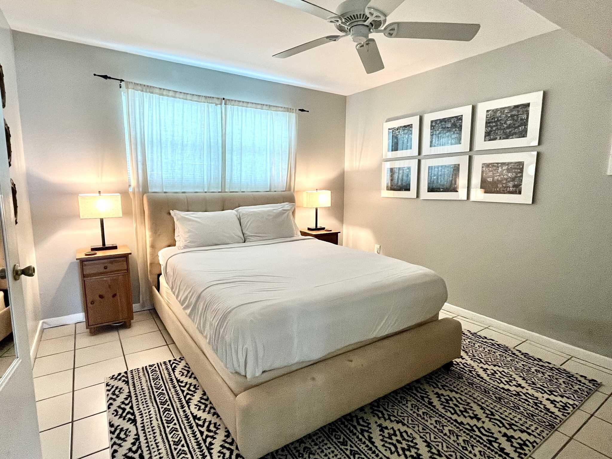 A bedroom with a bed, two bedside tables, a ceiling fan, a window with curtains, and a wall with framed pictures.