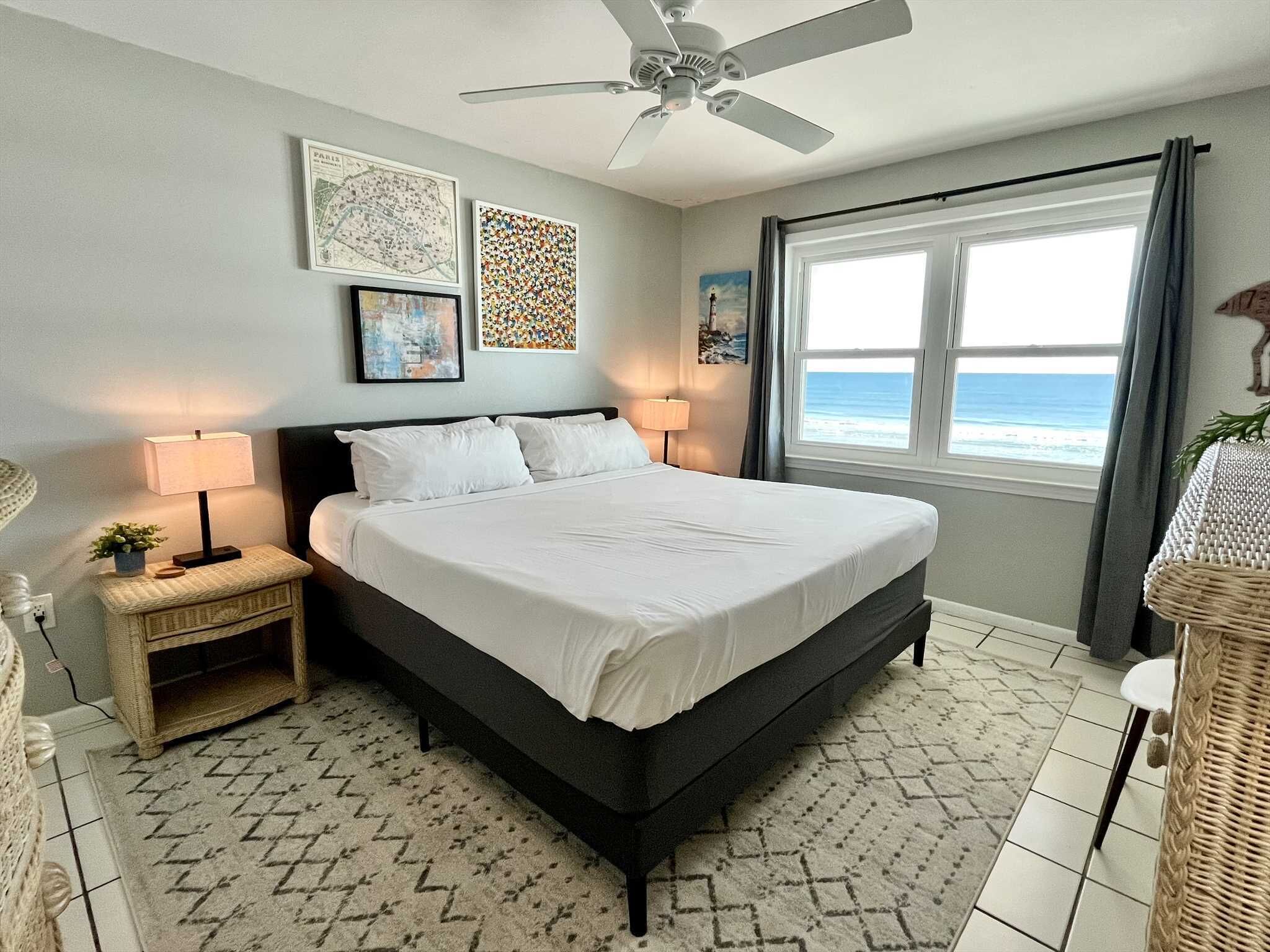 A bedroom with a bed, two bedside tables, a ceiling fan, a window with a view of the ocean, and decorative wall art.