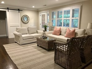 Een woonkamer met een beige bank, twee fauteuils, een salontafel en een groot raam met zonwering.