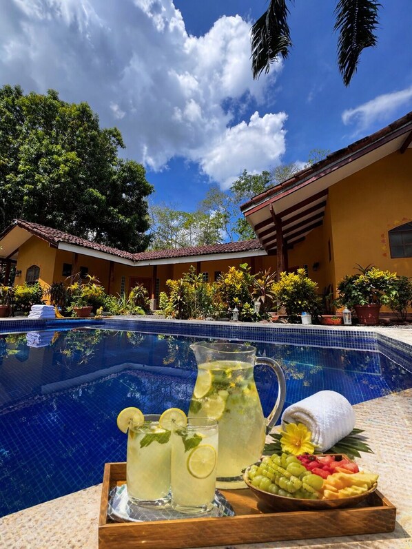 Ein Poolbereich mit klarem, blauem Pool, einer Serviertraufe mit Getränken und Früchten sowie einem Gebäude mit einem Ziegeldach im Hintergrund.