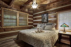 Quarto de cabana de madeira com cama, duas janelas com persianas, ventilador de teto, quadro emoldurado e abajur.