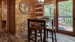 Ein Holzblockhaus-Interieur mit Esstisch und Barhocker, einer Wanddekoration mit Baum-Motiv und Blick ins Freie durch ein großes Fenster.
