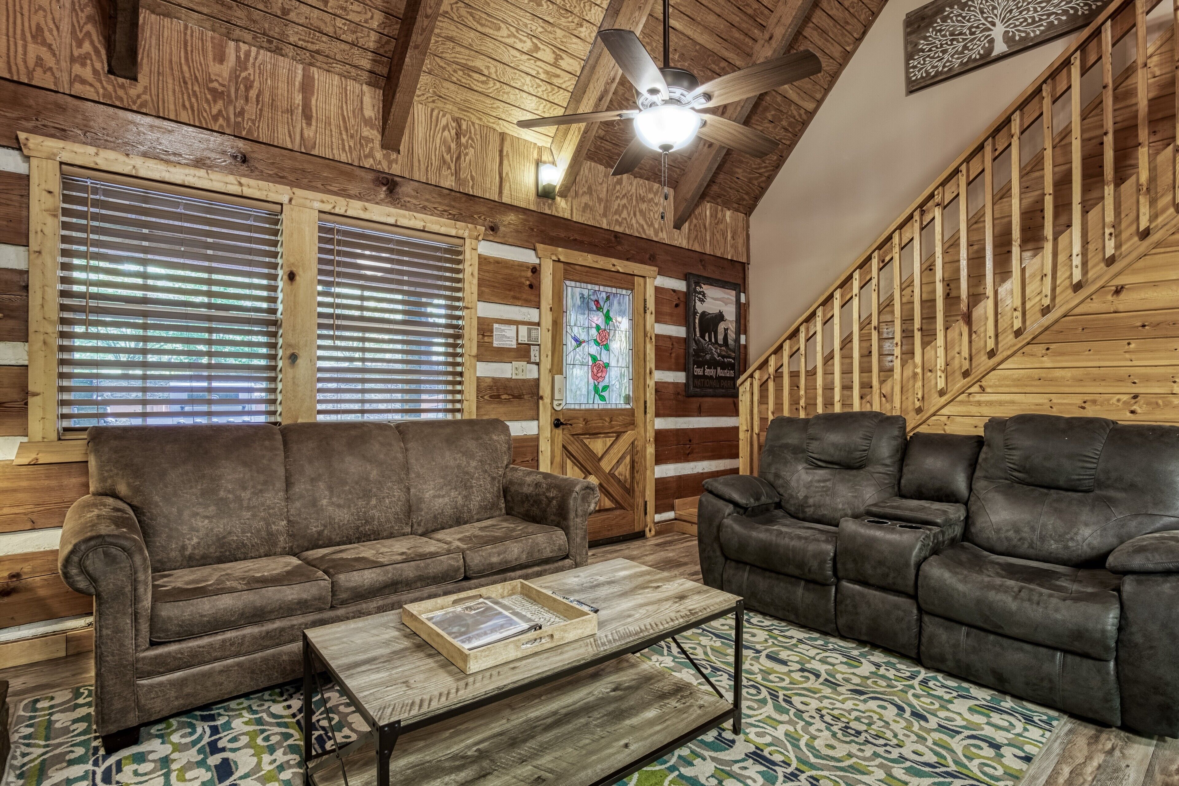 A cozy cabin interior with two sofas, a wooden coffee table, a ceiling fan, and a staircase.