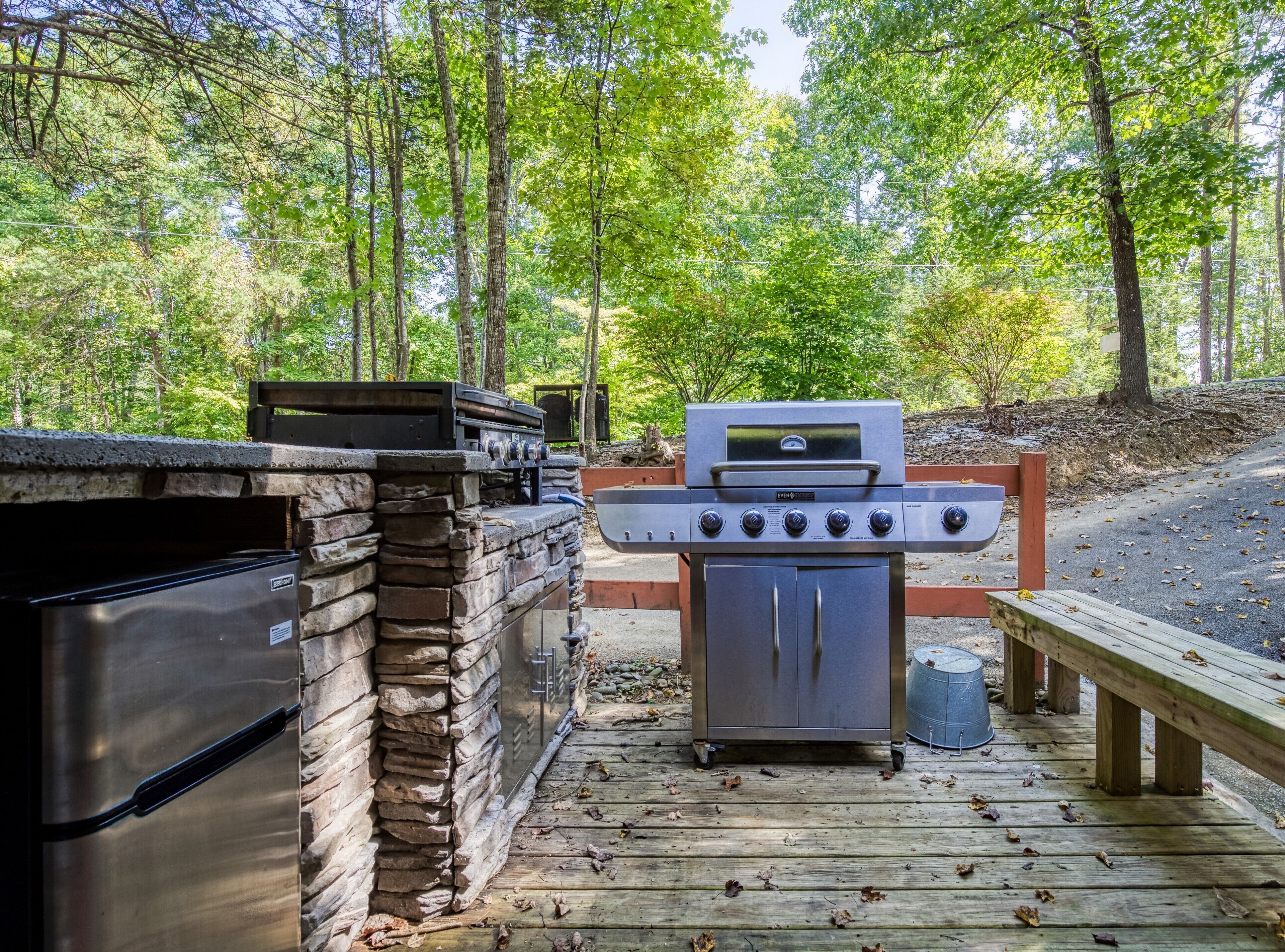 An outdoor kitchen with a built-in grill and a stainless steel refrigerator, set on a wooden deck surrounded by trees.