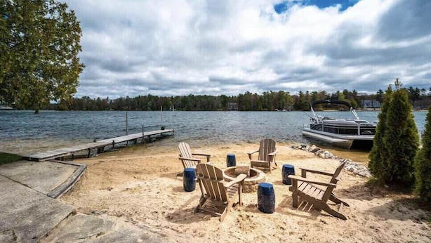 A lakeside deck with Adirondack chairs and a fire pit, overlooking a calm lake with a dock and boat.