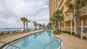 A swimming pool with a curved design, surrounded by lounge chairs and palm trees, adjacent to a building with balconies. - L👀K!!! ⭐️ TOP RATED ⭐️ DIRECT BEACHFRONT Condo at Calypso + FREE Beach Chairs !!! (Panama City Beach)
