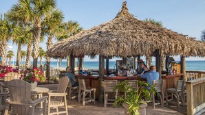 A beachside bar with wooden chairs and tables, a thatched roof, and a view of the ocean. - L👀K!!! ⭐️ TOP RATED ⭐️ DIRECT BEACHFRONT Condo at Calypso + FREE Beach Chairs !!! (Panama City Beach)