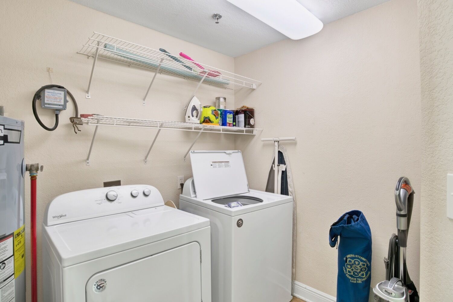 Une buanderie équipée de deux lave-linge et deux sèche-linge à chargement frontal, une étagère métallique au-dessus pour ranger les produits de nettoyage, et un aspirateur sur le côté droit.