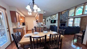A dining area with a wooden table and chairs, a kitchen in the background, and a stone fireplace.