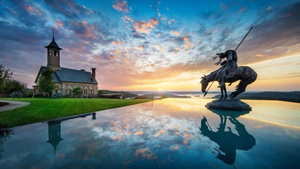 A church with a tower, a statue of a horse, and a calm reflective water surface.