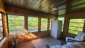 A cozy cabin interior with wooden walls, a large window overlooking greenery, a refrigerator, wicker furniture, and a potted plant. - River Ranch Rental - Cabin in the woods--fishing, relaxation, wildlife, river (Irons)