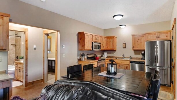 A modern kitchen with wooden cabinets, stainless steel appliances, a dining table with chairs, and a visible bathroom with a shower.