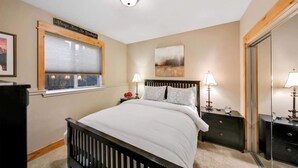 A bedroom with a bed, bedside tables, a window with blinds, a painting on the wall, and a mirror.