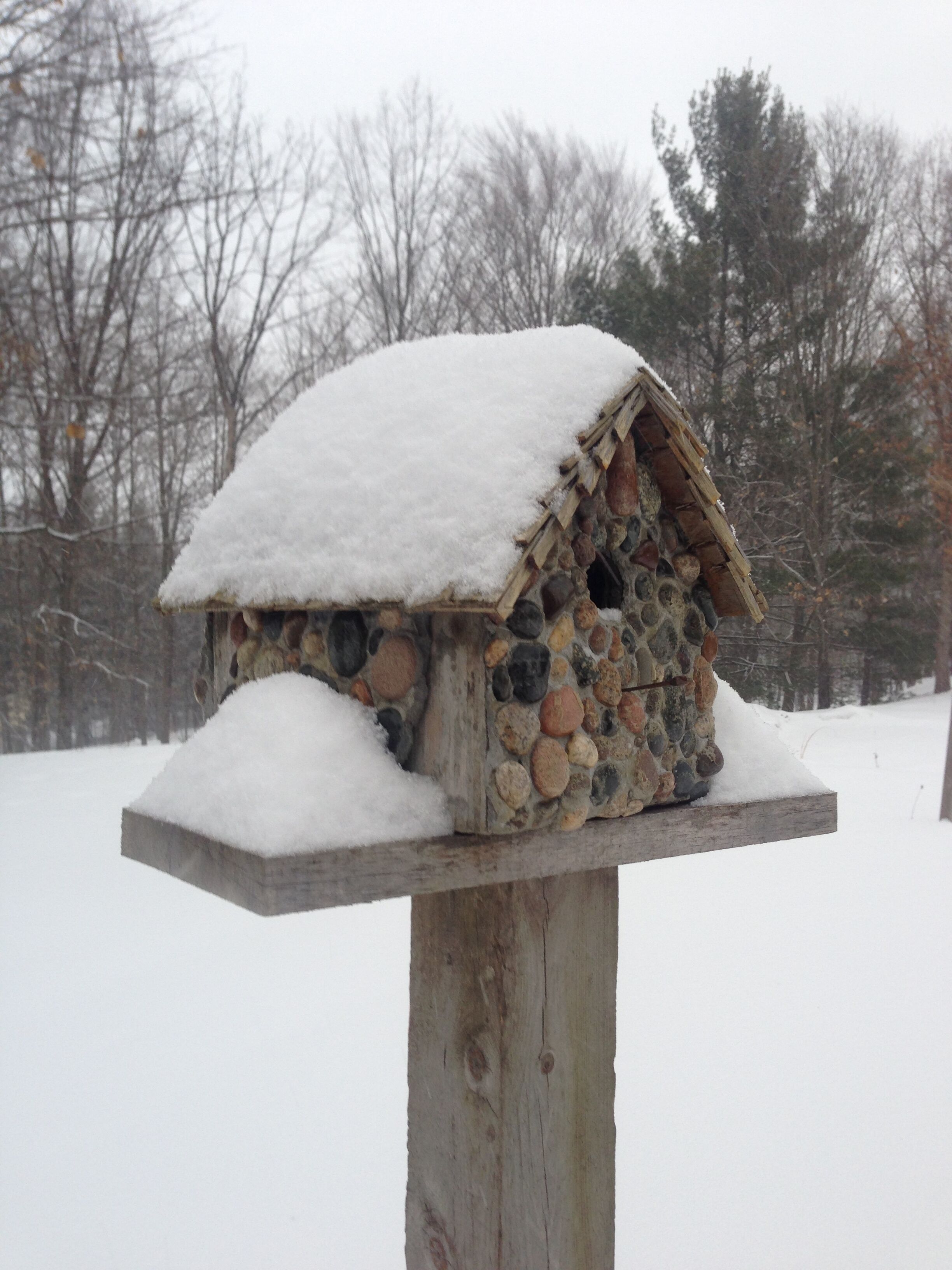 A stone birdhouse with a snow-covered roof, mounted on a wooden post.