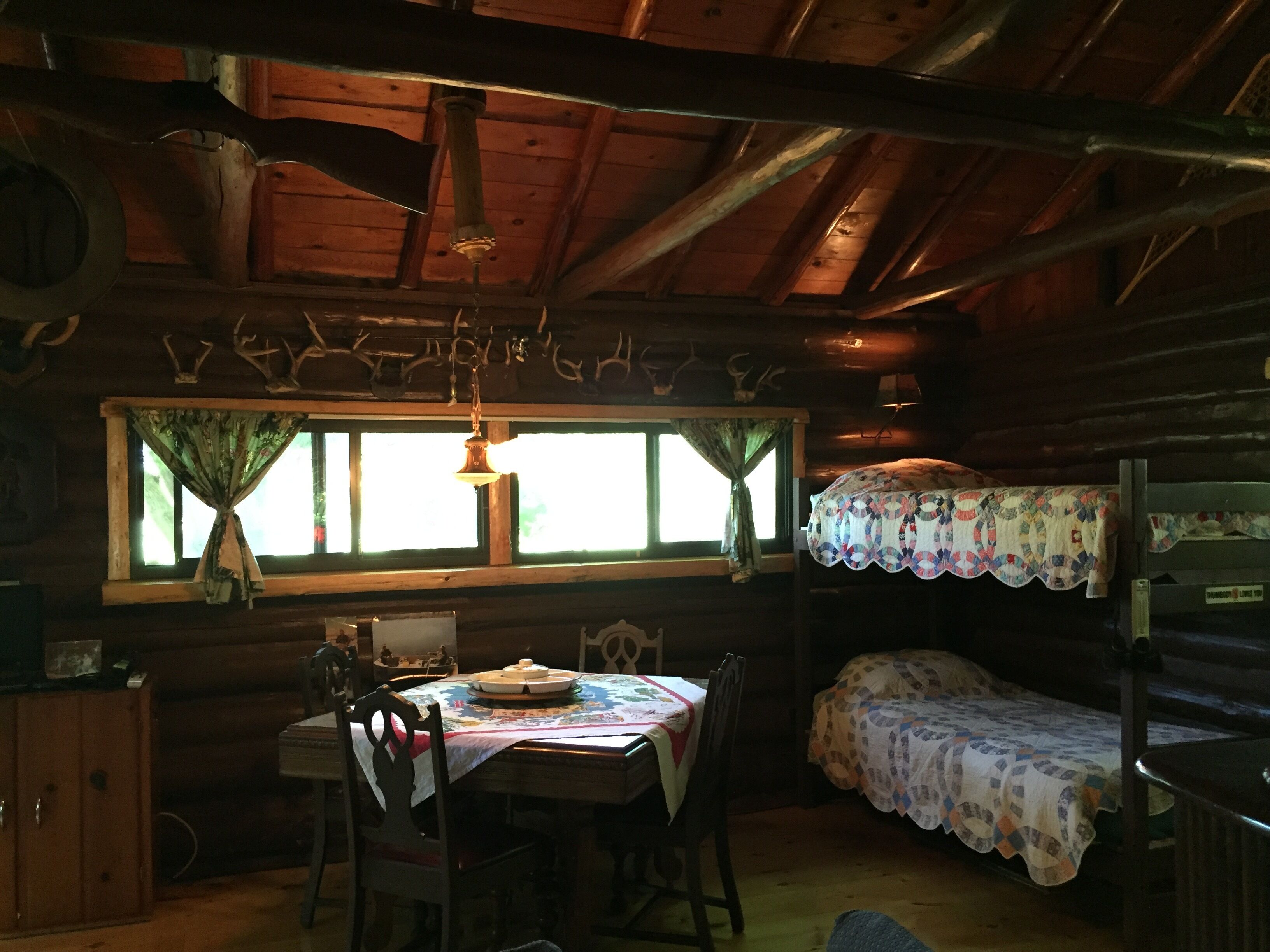 A rustic cabin interior with a wooden dining table, chairs, a bunk bed with a quilt, and a window with curtains.