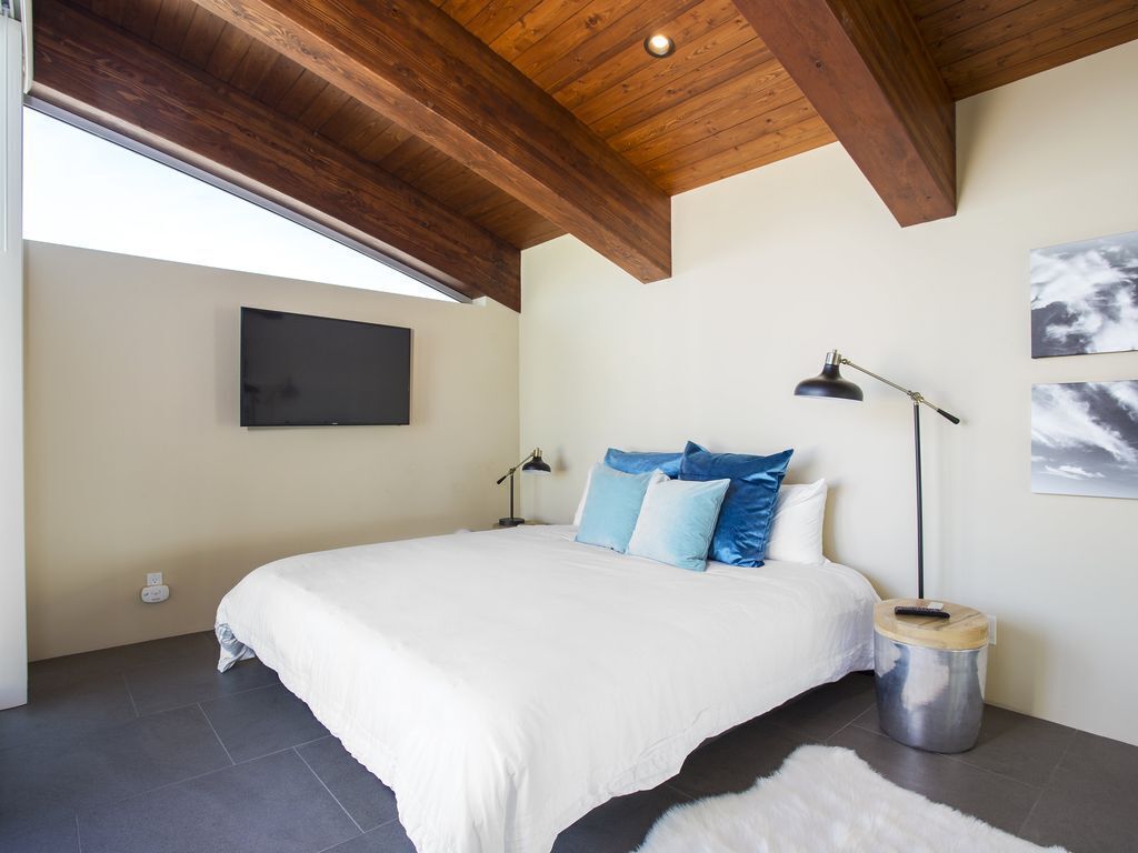 A bedroom with a large bed, a ceiling fan, a wall-mounted TV, and a small bedside table with a lamp.