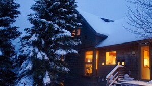 Une cabane en rondins, dont le toit est recouvert de neige et l’extérieur est en bois. On y voit plusieurs fenêtres illuminées, ainsi qu’un escalier menant à l’entrée.