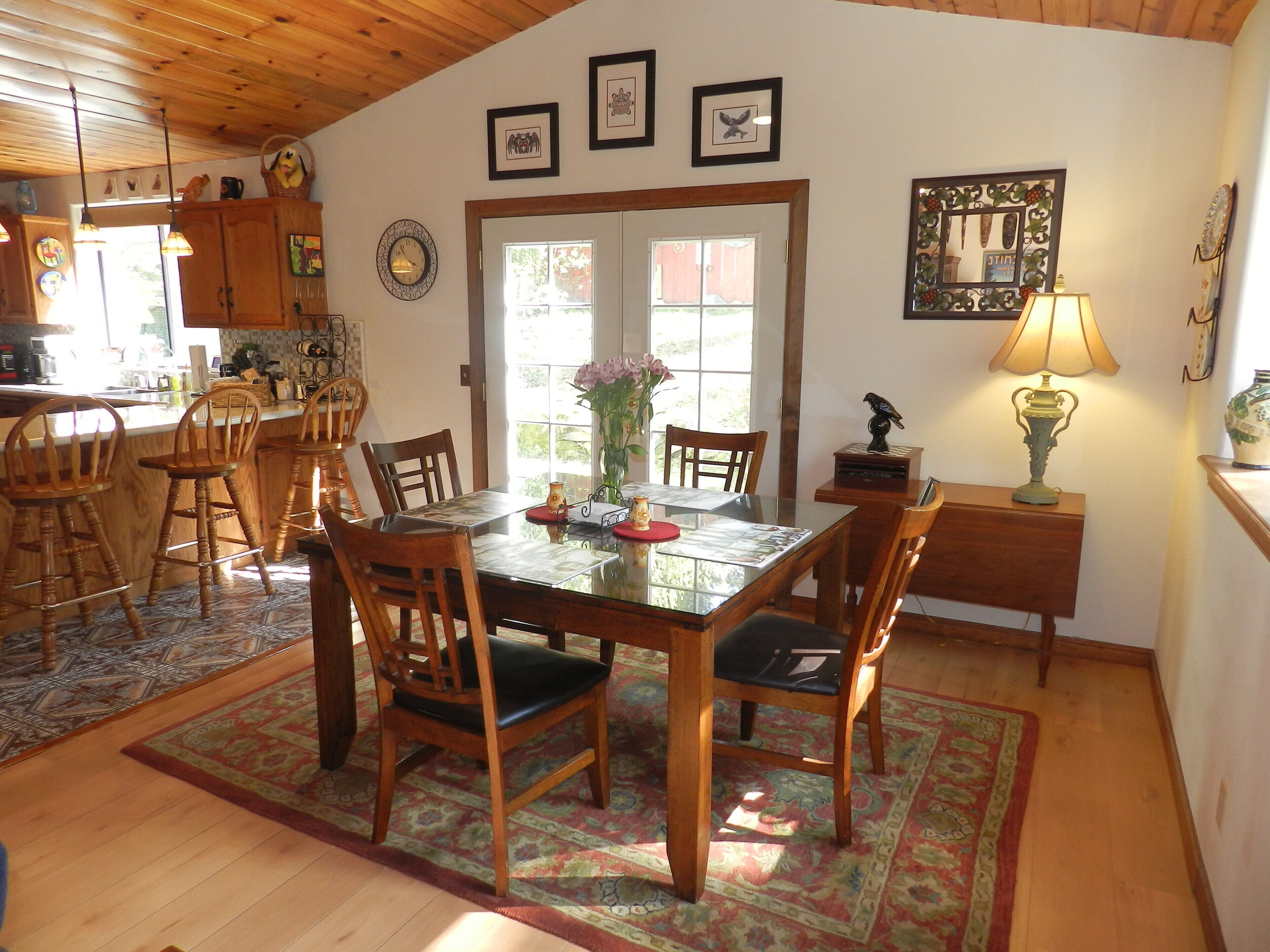 A dining area with a wooden table, chairs, and a rug. There is a window with a view of greenery outside.