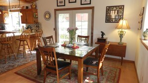 A dining area with a wooden table, chairs, and a rug. There is a window with a view of greenery outside. - Beautiful Creekside Home in Mariposa Pines Close to Yosemite.  No cleaning fee.  (Mariposa)