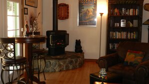 A cozy living room with a wood-burning stove, a bookshelf, and a poster of Yosemite Glacier Point Hotel. - Beautiful Creekside Home in Mariposa Pines Close to Yosemite.  No cleaning fee.  (Mariposa)