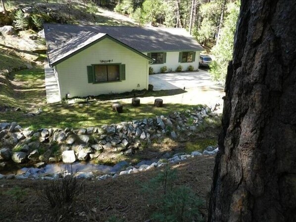 A small, single-story building with a sloped roof, surrounded by a stone wall and a grassy area with a small pond.