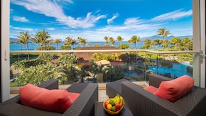 A balcony with wicker furniture, a fruit bowl, and a view of a pool and tropical landscape. - Oceanfront Views Above Dukes - Wrap Around Lanai - Outdoor Kitchen 🌺 Honua Kai H351 (Lahaina)