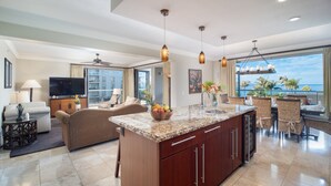 A spacious living area with a large TV, comfortable seating, a kitchen island, and a balcony with a view of the ocean. - Oceanfront Views Above Dukes - Wrap Around Lanai - Outdoor Kitchen 🌺 Honua Kai H351 (Lahaina)
