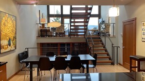 Ein geräumiger Essbereich mit einem langen Tisch und Stühlen, einer Treppe, die zu einem Balkon führt, und einem großen Fenster mit Blick ins Freie.