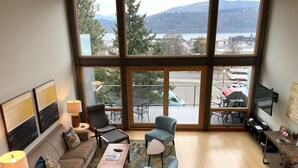 Ein großzügiger Wohnraum mit einem großen Fenster und Blick auf einen See und Berge. Es gibt ein Sofa, zwei Sessel, einen Couchtisch und einen an der Wand montierten Fernseher.