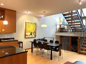 A modern dining area with a wooden table and chairs, a painting on the wall, and a staircase leading to an upper level.