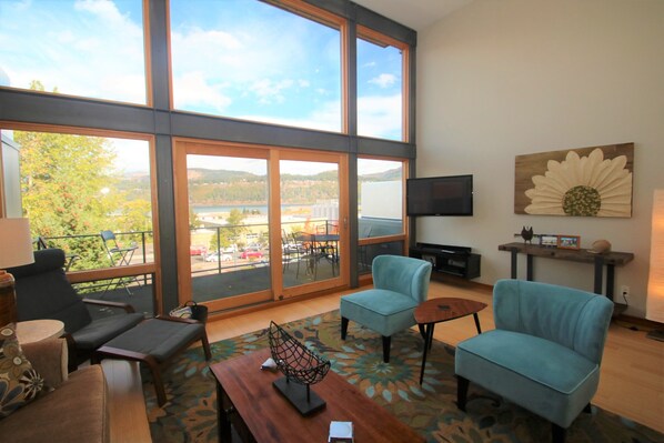 A spacious living area with large windows, a flat-screen TV, and a view of a lake and hills.