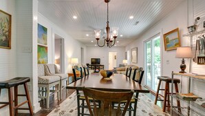 A dining area with a wooden table, chairs, and a chandelier. There are paintings on the walls and a television in the background. - Charming 125 Year Old Sewanee Home on Campus- Walk Everywhere! (Sewanee)