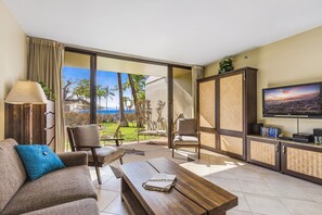 Ein großzügiger Wohnbereich mit einer Couch, Sesseln, einem Couchtisch und einem Fernseher. Das Zimmer verfügt über eine große Schiebeglastür, die auf einen Balkon mit Blick auf Palmen und das Meer führt.