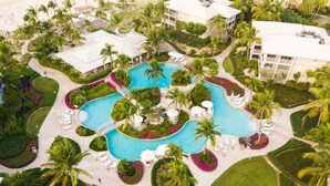 An aerial view of a resort with a central swimming pool, surrounded by palm trees, lounge chairs, and well-manicured gardens. - Updated Stunning Ocean Club Plaza luxury family spacious top floor apartment  (Providenciales)