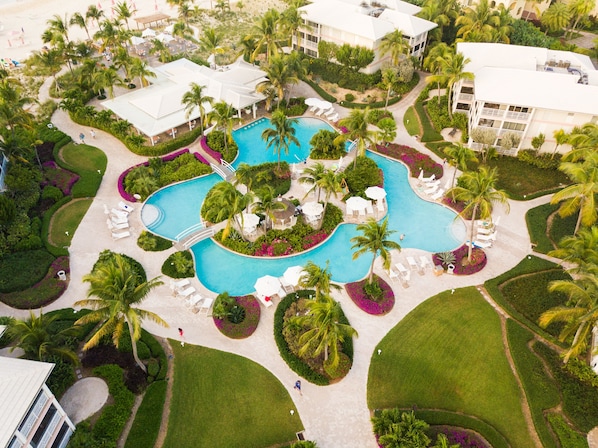 An aerial view of a resort with a central swimming pool, surrounded by palm trees, lounge chairs, and well-manicured gardens.
