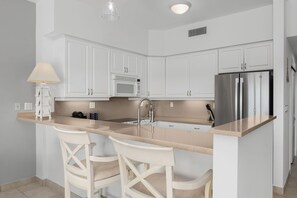 Una cocina moderna con armarios blancos, un refrigerador de acero inoxidable, un microondas y una barra con dos taburetes blancos.