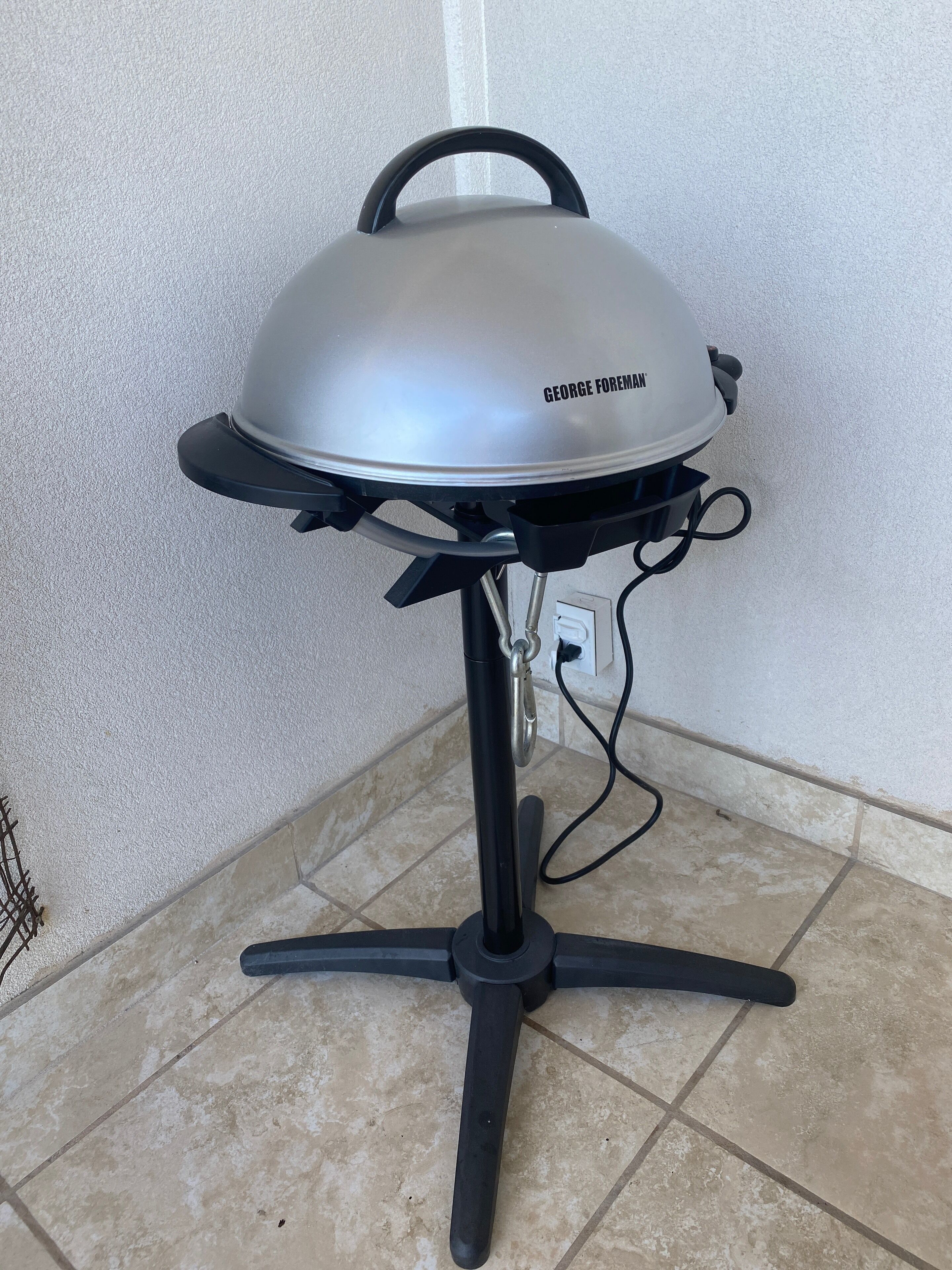 Ein tragbarer Grill mit schwarzem Gestell und silbernem Deckel, beschriftet mit "GEORGE FOREMAN", steht auf einem gefliesten Boden vor einer weißen Wand.