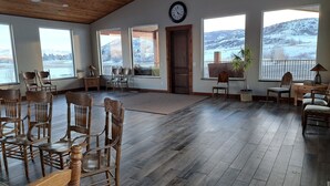 Ein geräumiges Zimmer mit Holzboden, großen Fenstern mit Blick auf eine verschneite Landschaft und mehreren Holzsesseln und -tischen.