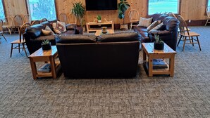 Ein gemütliches Wohnzimmer mit Ledersofas, Holztischen und Stühlen. An den Wänden sind aufgehängte Tierköpfe und ein großes Fenster mit Blick ins Freie.