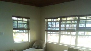 Ein Wohnzimmer mit zwei Sofas, einem Couchtisch und einem kleinen Beistelltisch. Es gibt Fenster mit Jalousien, und auf dem Couchtisch steht eine Pflanze.
