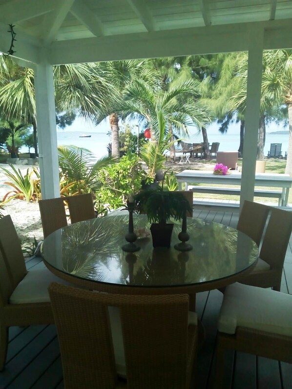 Un área de comedor al aire libre techada, con una mesa de vidrio y sillas de mimbre, rodeada de plantas tropicales y con vista a una playa donde se divisa un barco a lo lejos.